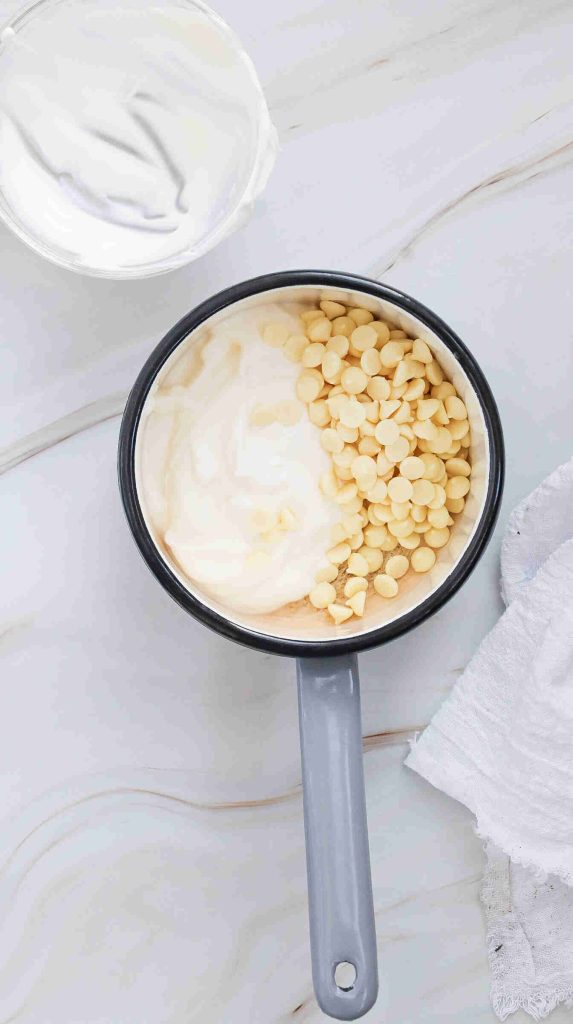 A saucepan with white chocolate chips and cream, perfect for making iced white chocolate mocha, sits on a marble countertop next to a bowl of cream and a white cloth napkin.