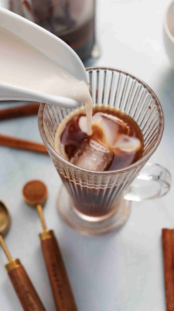 A glass cup of iced cinnamon almond milk latte sits on a table while milk is being poured in, creating swirling patterns. Nearby are cinnamon sticks and gold measuring spoons.