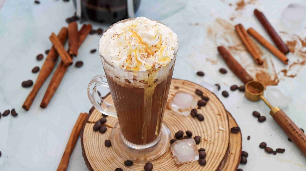 A glass mug of iced cinnamon almond milk latte topped with whipped cream and caramel drizzle sits on a wooden coaster, surrounded by coffee beans, cinnamon sticks, ice cubes, and a gold coffee scoop.