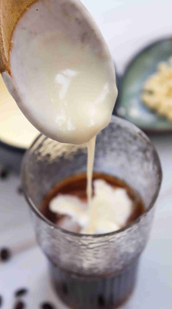A spoon pours creamy milk or cream into a glass of iced white chocolate mocha, creating swirling patterns in the dark liquid. A plate with food is blurred in the background.