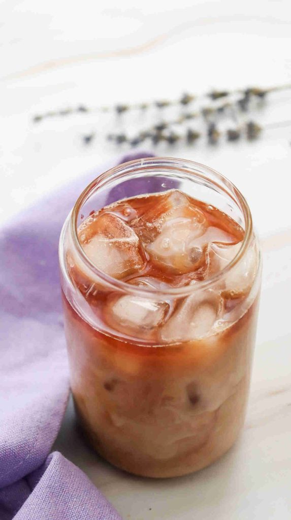 A glass jar of lavender iced coffee, with visible ice cubes and creamy swirls, sits on a white surface next to a lavender cloth and sprigs of dried lavender in the background.