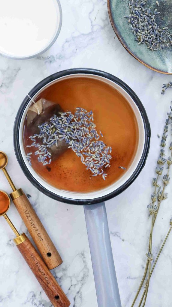 A saucepan filled with earl grey milk tea, a tea bag, and lavender buds sits on a marble surface. Surrounding it are a plate of lavender, a cup of milk, gold measuring spoons, and sprigs of dried lavender.