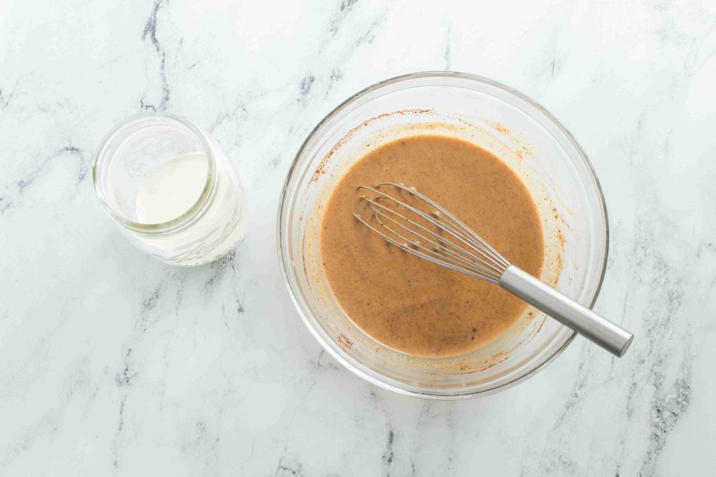 A glass bowl with a metal whisk mixing a brown batter sits on a marble surface next to a small glass jar filled with pumpkin spice coffee creamer.