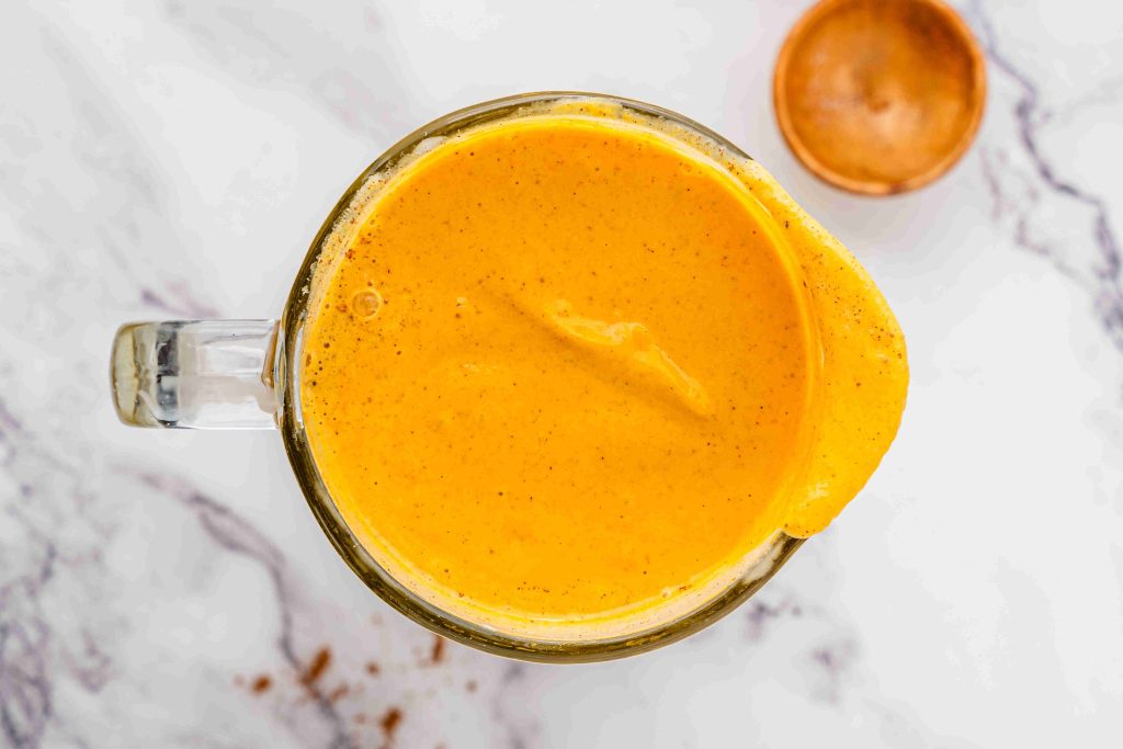 A glass blender viewed from above, filled with a creamy, bright orange pumpkin pie spice milkshake spilling slightly over the rim, set on a white marble surface with a small wooden bowl nearby.