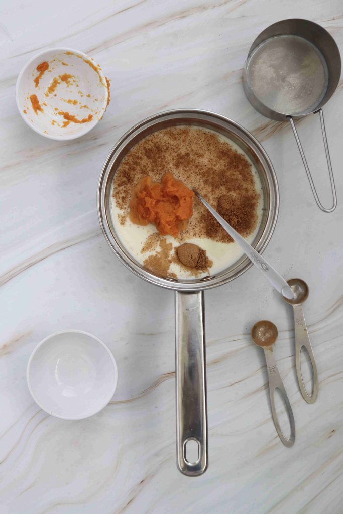 A saucepan filled with almond milk, canned pumpkin, brown sugar, and spices for a homemade pumpkin spice latte sits on a marble countertop beside two small white bowls, two measuring spoons, and a metal measuring cup.