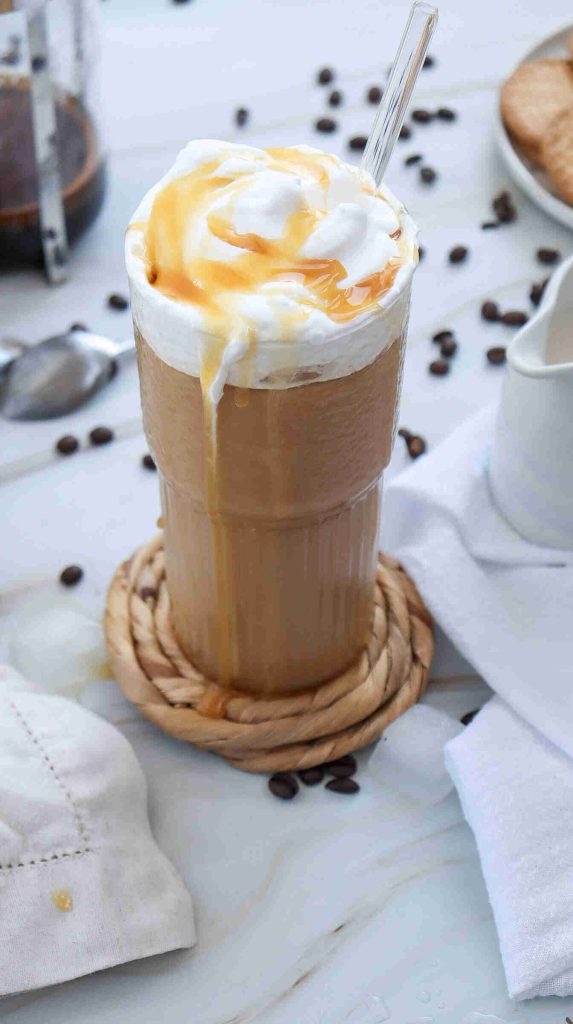 A tall glass of iced coffee topped with whipped cream and caramel drizzle sits on a woven coaster, hinting at how to make caramel iced coffee at home, with a clear straw, scattered coffee beans, and white napkins on a light surface.