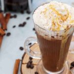 A glass mug filled with an iced cinnamon almond milk latte, topped with whipped cream and caramel drizzle. Coffee beans, cinnamon sticks, and ice cubes are scattered on a wooden surface around the mug.