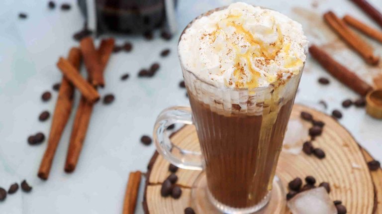A glass mug filled with an iced cinnamon almond milk latte, topped with whipped cream and caramel drizzle. Coffee beans, cinnamon sticks, and ice cubes are scattered on a wooden surface around the mug.