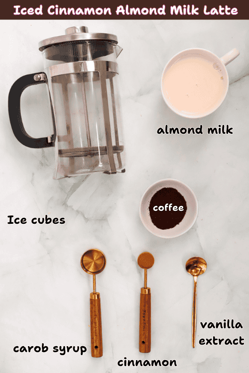 Overhead view of ingredients for an iced cinnamon almond milk latte: a French press, a cup of almond milk, a bowl of coffee, ice cubes, and three gold measuring spoons with carob syrup, cinnamon, and vanilla extract.