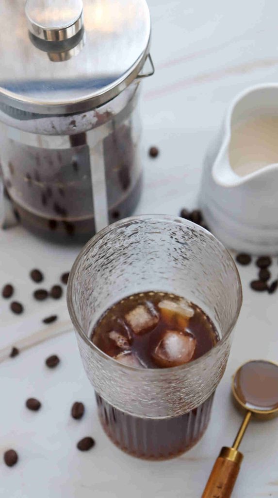 A glass of iced coffee with ice cubes sits on a marble surface, surrounded by coffee beans—perfect inspiration for anyone curious about how to make caramel iced coffee at home. Behind the glass is a French press and a small white creamer. A spoon rests nearby.