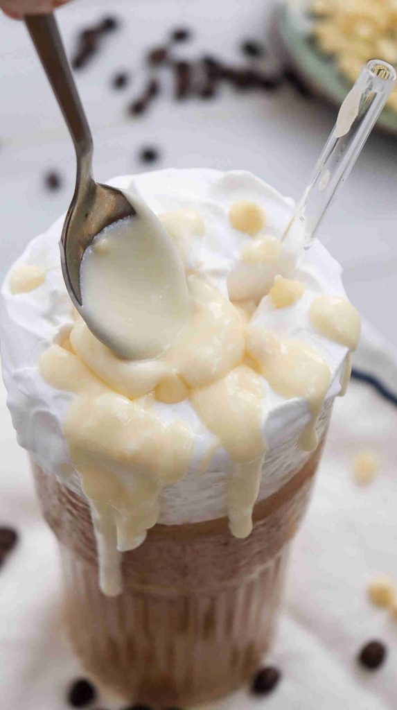 A glass of iced white chocolate mocha topped with whipped cream is being drizzled with thick, creamy condensed milk from a spoon. A clear straw is inserted into the drink, and some coffee beans are scattered around the glass.