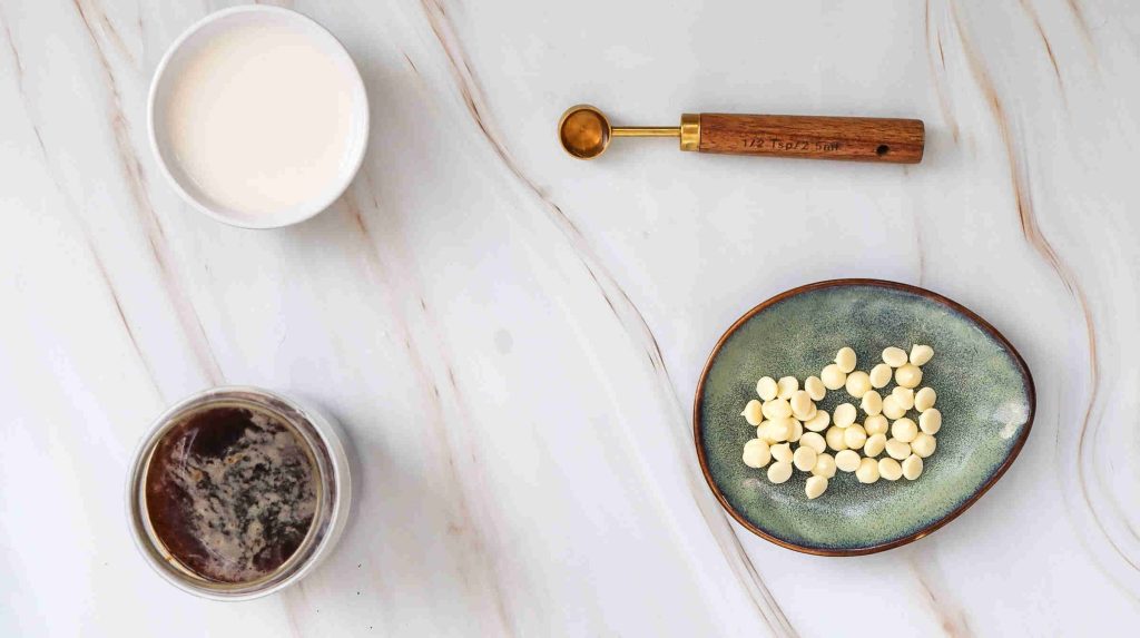 A small bowl of cream, a jar with brown liquid for an iced white chocolate mocha, a teaspoon, and a plate with white chocolate chips are arranged on a white marble surface.