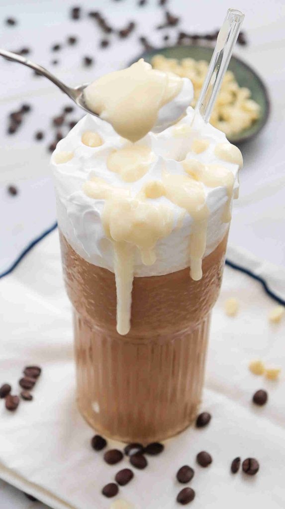 A glass of iced white chocolate mocha topped with whipped cream and drizzled with a creamy sauce, with a spoon hovering above and coffee beans scattered around on a white cloth.