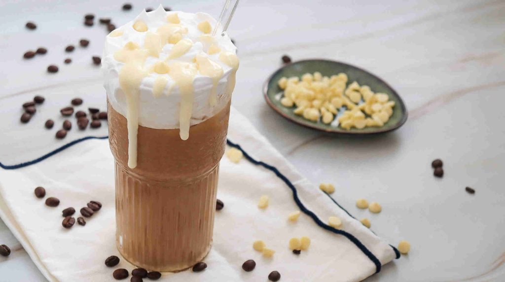 Iced white chocolate mocha topped with whipped cream and white chocolate chips, served in a tall glass with a straw. Coffee beans and white chocolate chips are scattered on a napkin, and a dish of chips is in the background.