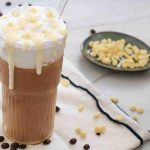 Iced white chocolate mocha topped with whipped cream and white chocolate chips, served in a tall glass with a straw. Coffee beans and white chocolate chips are scattered on a napkin, and a dish of chips is in the background.