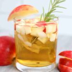 A glass of golden Apple Cider Whiskey Cocktail garnished with diced red apples, a sprig of rosemary, and an apple slice. Whole and sliced apples rest on a light wooden surface beside the glass.