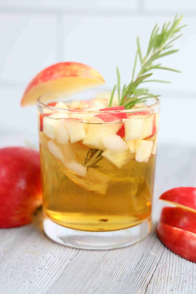 A glass of golden Apple Cider Whiskey Cocktail garnished with diced red apples, a sprig of rosemary, and an apple slice. Whole and sliced apples rest on a light wooden surface beside the glass.