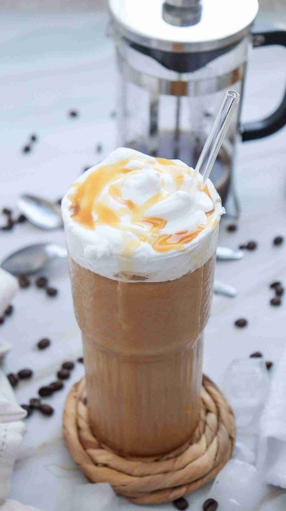 A glass of iced coffee topped with whipped cream and caramel drizzle, served with a clear straw. Coffee beans, spoons, and a French press blur in the background—perfect if you’re curious about how to make caramel iced coffee at home.