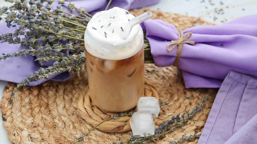 A lavender iced coffee topped with whipped cream and lavender petals sits on a woven mat, surrounded by sprigs of lavender, two ice cubes, and folded purple napkins tied with twine.