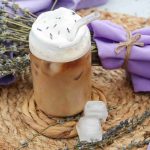 A lavender iced coffee topped with whipped cream and lavender petals sits on a woven mat, surrounded by sprigs of lavender, two ice cubes, and folded purple napkins tied with twine.