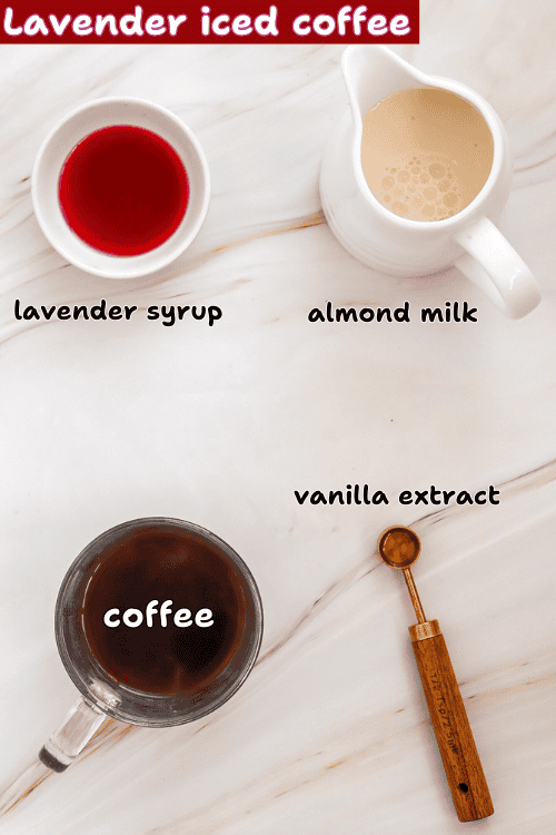 Overhead view of ingredients for lavender iced coffee: a small bowl of lavender syrup, a small pitcher of almond milk, a mug of coffee, and a measuring spoon with vanilla extract are arranged on a marble surface for this refreshing drink.