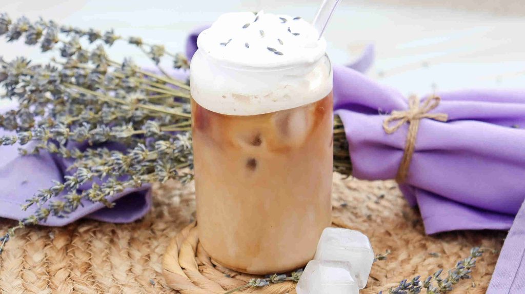 A glass of lavender iced coffee with whipped cream and lavender buds on top, placed on a wicker mat beside a bundle of lavender tied with purple cloth and two ice cubes.