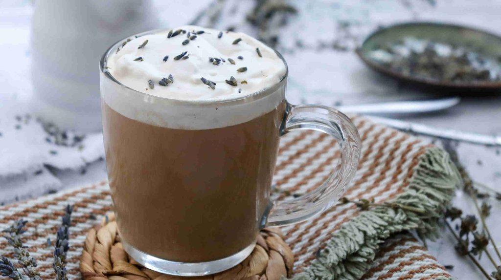 A glass mug of earl grey milk tea topped with creamy foam and sprinkled with lavender buds, placed on a woven coaster with a striped cloth and lavender sprigs in the background.