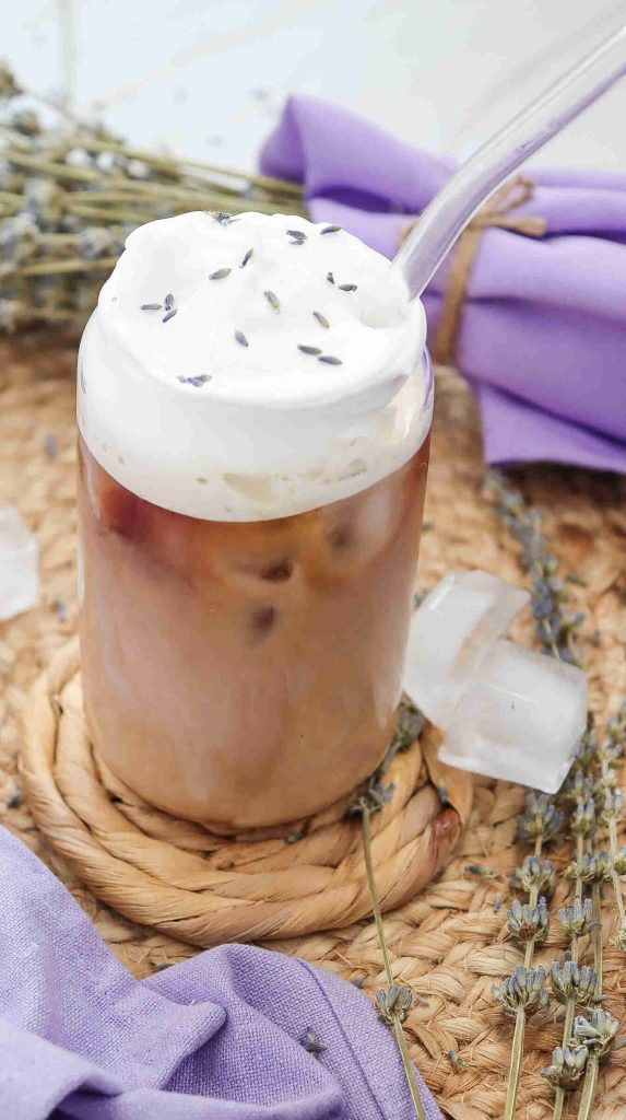A glass of lavender iced coffee topped with whipped cream and lavender buds, served with a clear straw. Ice cubes, lavender sprigs, and a purple napkin are arranged on a woven mat in the background.