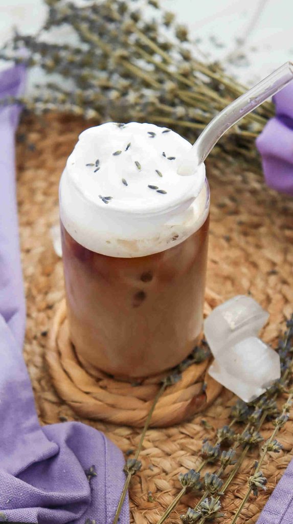 A glass of lavender iced coffee topped with thick whipped cream and garnished with lavender buds sits on a woven mat, with a glass straw, ice cubes, and sprigs of dried lavender in the background.