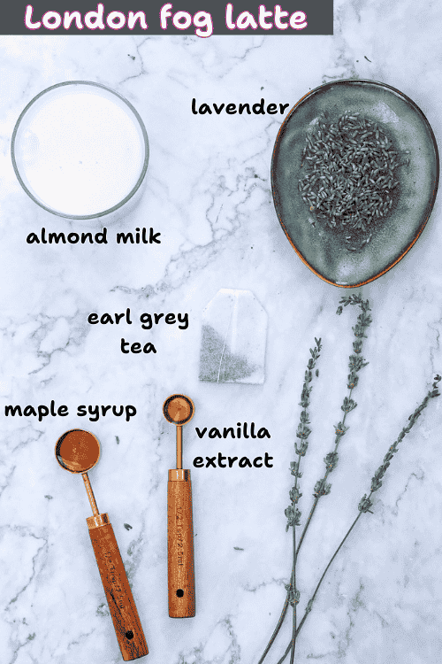 Ingredients for a London fog latte on a marble surface: a glass of almond milk, Earl Grey tea bag for the perfect earl grey milk tea blend, a dish of lavender, wooden spoons with maple syrup and vanilla extract, and sprigs of lavender.