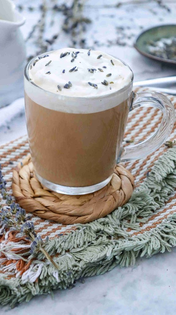 A clear glass mug of earl grey milk tea latte with a layer of frothy milk topped with lavender buds sits on a woven coaster atop textured, fringed fabric. Lavender sprigs and a green plate adorn the background.