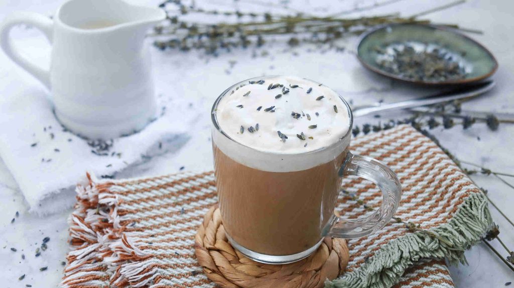 A glass mug of frothy lavender latte topped with dried lavender buds sits on a woven coaster and striped towel, alongside a small pitcher, plate, loose lavender, and hints of earl grey milk tea in the background.