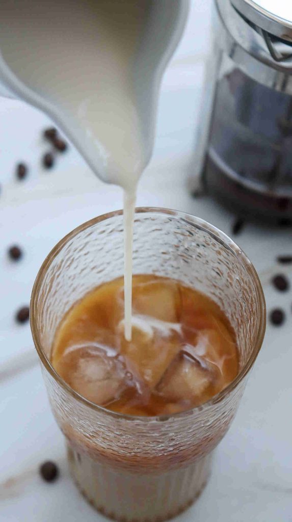 A hand pours creamy milk into a glass of iced coffee with ice cubes, surrounded by scattered coffee beans and a French press—perfect inspiration for how to make caramel iced coffee at home.