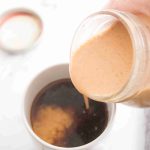 A hand pours creamy, light brown pumpkin spice coffee creamer from a glass jar into a mug of black coffee, creating a swirl. The jar lid is visible in the background on a white surface.