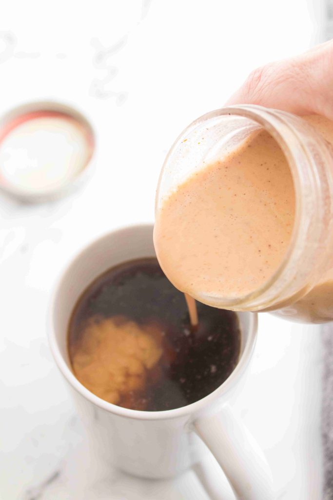 A hand pours creamy, light brown pumpkin spice coffee creamer from a glass jar into a mug of black coffee, creating a swirl. The jar lid is visible in the background on a white surface.