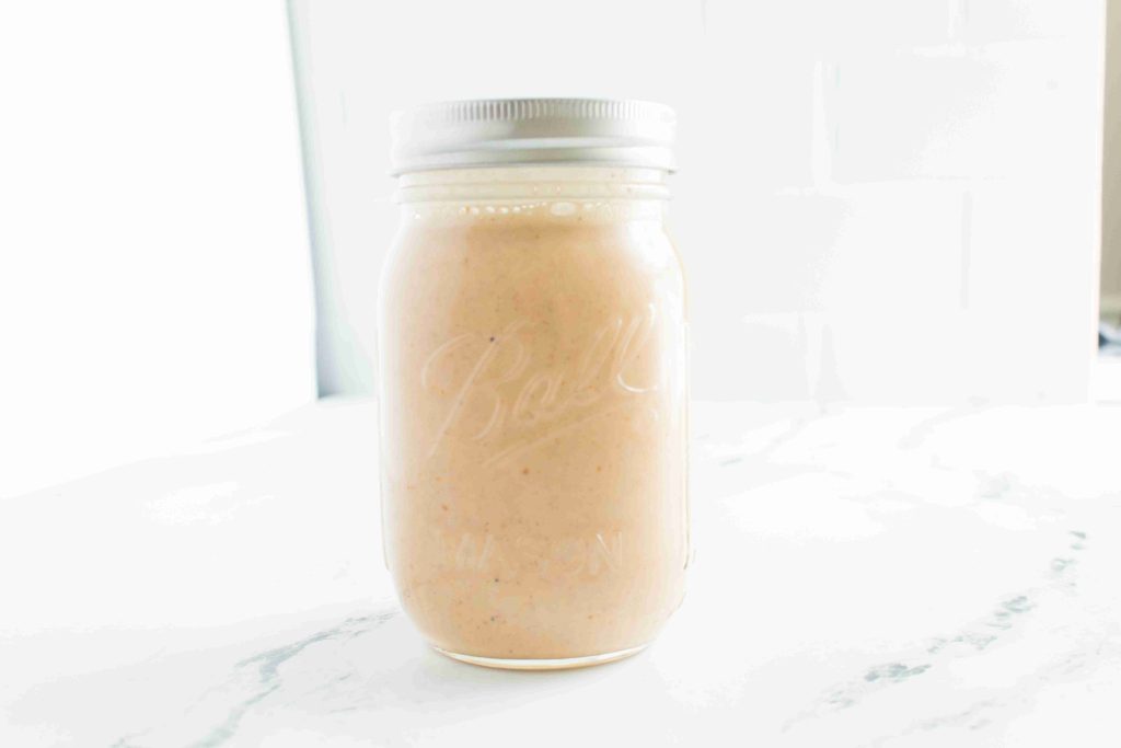 A glass mason jar with a metal lid is filled with creamy, light pink pumpkin spice coffee creamer, sitting on a white marble surface with a bright, softly lit background.