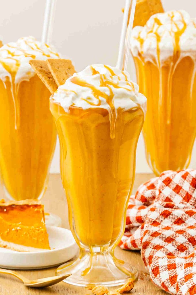 Three tall glasses filled with creamy pumpkin pie spice milkshake, topped with whipped cream, caramel drizzle, and a graham cracker. A slice of pumpkin pie and a red-checked napkin are visible nearby.