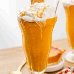 A tall glass filled with a creamy pumpkin pie spice milkshake, topped with whipped cream, caramel drizzle, and a piece of graham cracker. A glass straw is inserted, with a slice of pie and a spoon in the background.