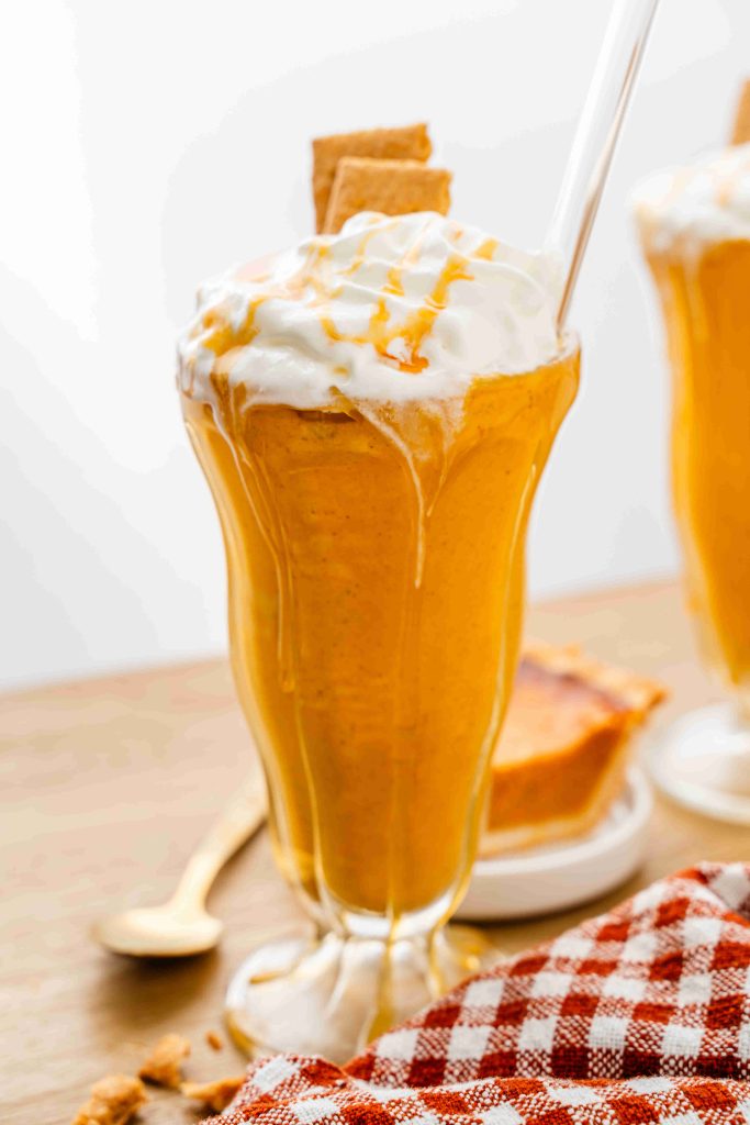 A tall glass filled with a creamy pumpkin pie spice milkshake, topped with whipped cream, caramel drizzle, and a piece of graham cracker. A glass straw is inserted, with a slice of pie and a spoon in the background.