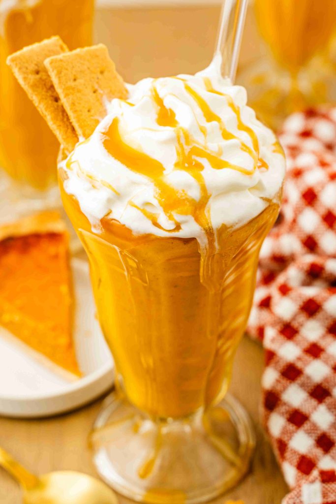 A tall glass filled with pumpkin pie spice milkshake, topped with whipped cream and caramel drizzle, garnished with two graham crackers. A slice of pumpkin pie and a red-checkered cloth are in the background.