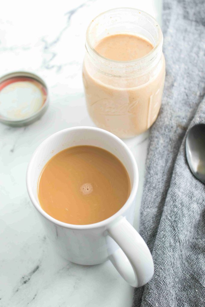 A white mug filled with light brown coffee sits on a white surface next to an open mason jar of pumpkin spice coffee creamer, a metal spoon, a lid, and a gray cloth napkin.