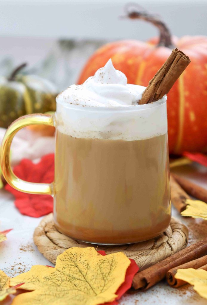 A pumpkin spice latte with almond milk sits in a glass mug, topped with whipped cream and a cinnamon stick, surrounded by autumn leaves, cinnamon sticks, and pumpkins in a cozy fall setting.