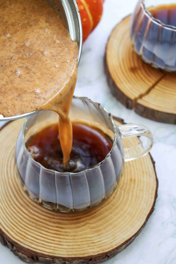 A creamy, spiced dairy free pumpkin spice latte mixture is being poured from a saucepan into a glass mug of coffee, resting on a wooden coaster. Another filled mug sits nearby, also on a wooden coaster.