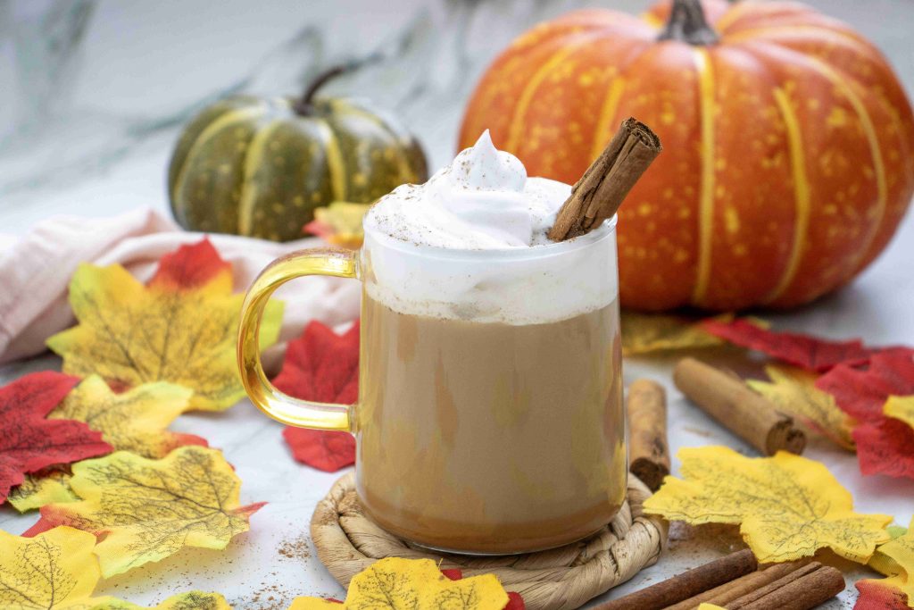 A glass mug of pumpkin spice latte with almond milk, topped with whipped cream and a cinnamon stick, sits among colorful autumn leaves, cinnamon sticks, and two pumpkins in the background.