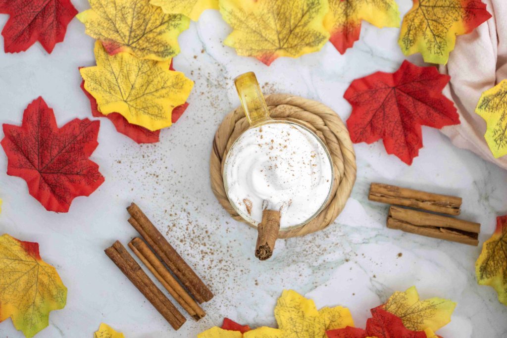 A mug of pumpkin spice latte with almond milk, topped with whipped cream and a cinnamon stick, is surrounded by artificial red and yellow autumn leaves and cinnamon sticks on a marble surface.