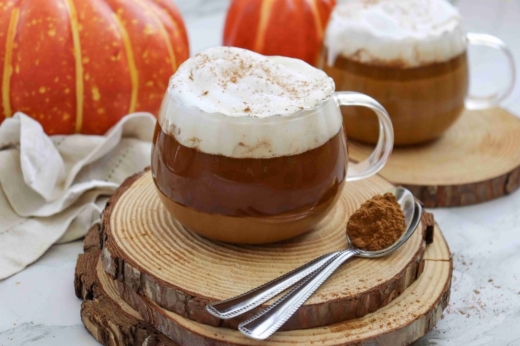 A glass mug of dairy free pumpkin spice latte with whipped cream sits on wooden coasters, next to two spoons with cinnamon powder. Another mug and decorative pumpkins are in the background.