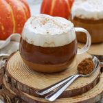 A glass mug of dairy free pumpkin spice latte with whipped cream sits on wooden coasters, next to two spoons with cinnamon powder. Another mug and decorative pumpkins are in the background.