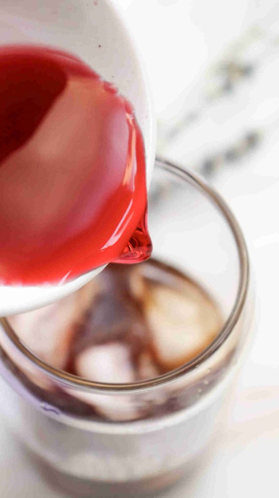 A close-up of red liquid being poured from a small white pitcher into a glass filled with ice cubes, creating a vibrant twist on classic lavender iced coffee.