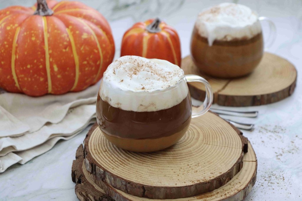 Two glass mugs of dairy free pumpkin spice latte topped with frothy whipped cream and a sprinkle of cinnamon sit on wooden coasters. Two pumpkins and napkins are in the background on a marble surface.