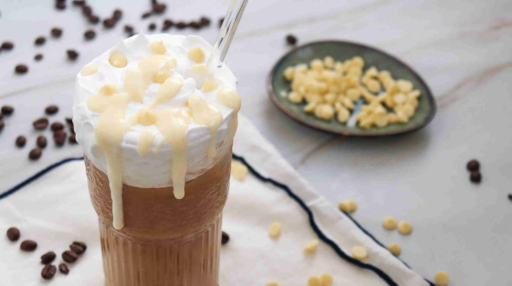 A glass of iced white chocolate mocha topped with whipped cream and white chocolate drizzle sits with a straw. Coffee beans and white chocolate chips are scattered nearby, with a small plate of white chocolate chips in the background.
